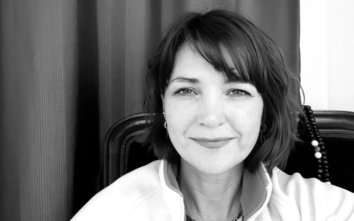 Black and white photo of Terryl Maddocks smiling, seated in her studio with mala beads on the chair behind her